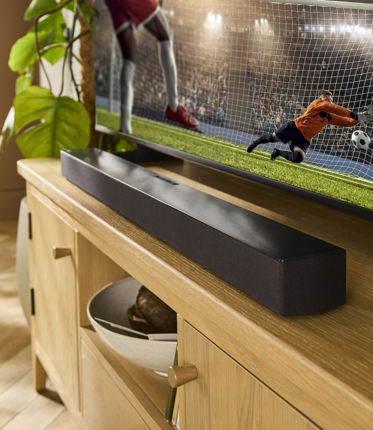 A black soundbar on a TV unit in front of a TV on displaying a football match in action.