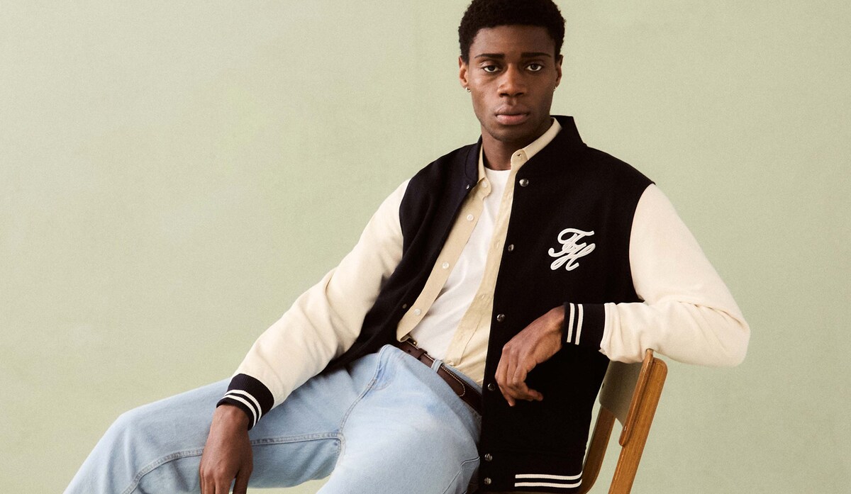 Model is wearing a Tommy Hilfiger Script Colourblock bomber jacket over a white t-shirt and cream shirt with a pair of light blue jeans.