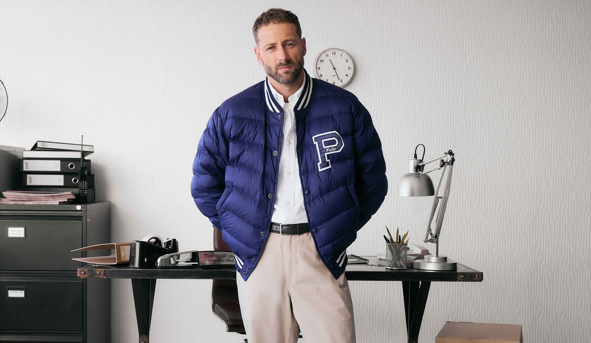 Model is wearing a navy Polo Ralph Lauren Logo bomber jacket over a white shirt and cream trousers.