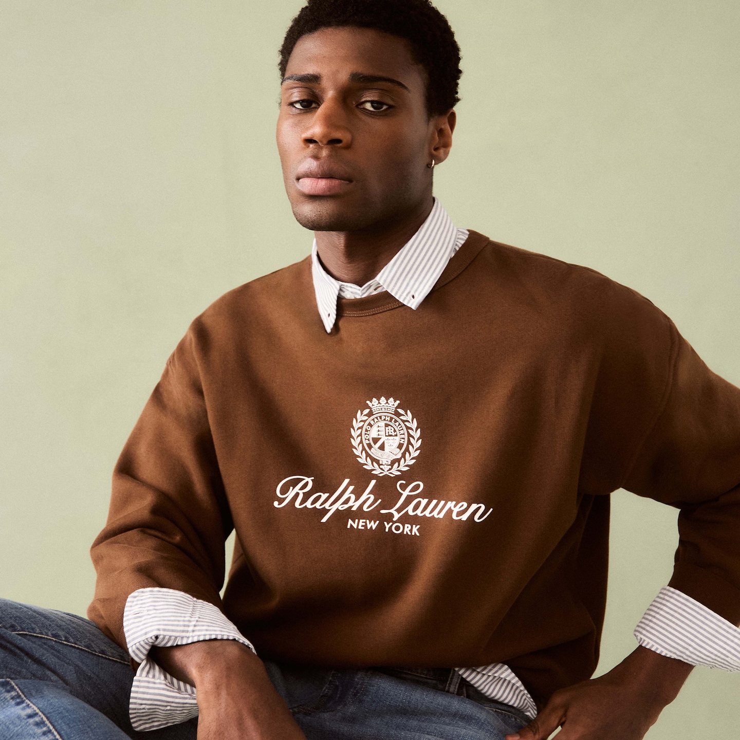 Model is wearing a brown Polo Ralph Lauren graphic crew neck sweatshirt with a stripe shirt underneath with a pair of jeans.