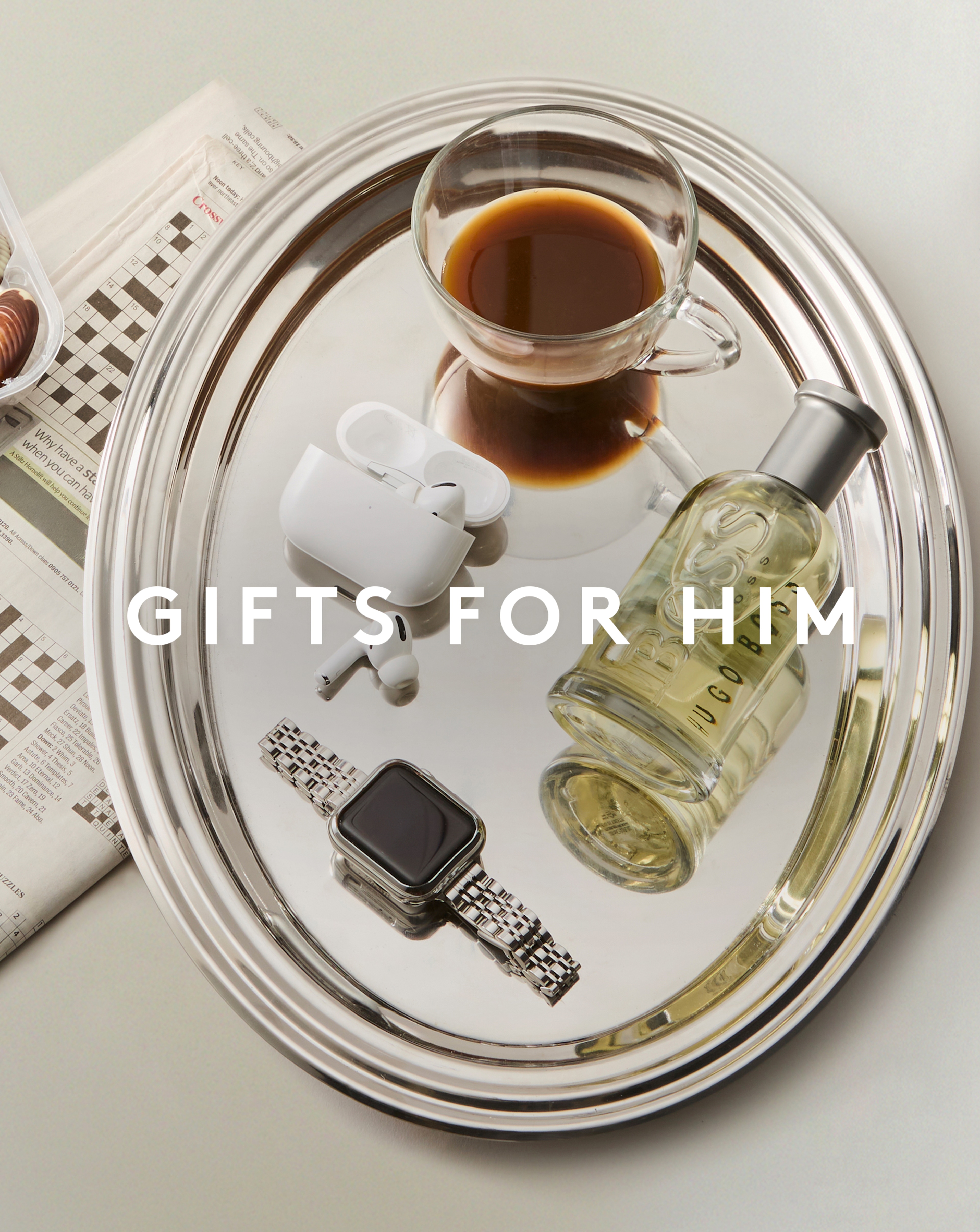 Gifts for him. A glass coffee cup on a silver tray with airpods, aftershave and a watch.