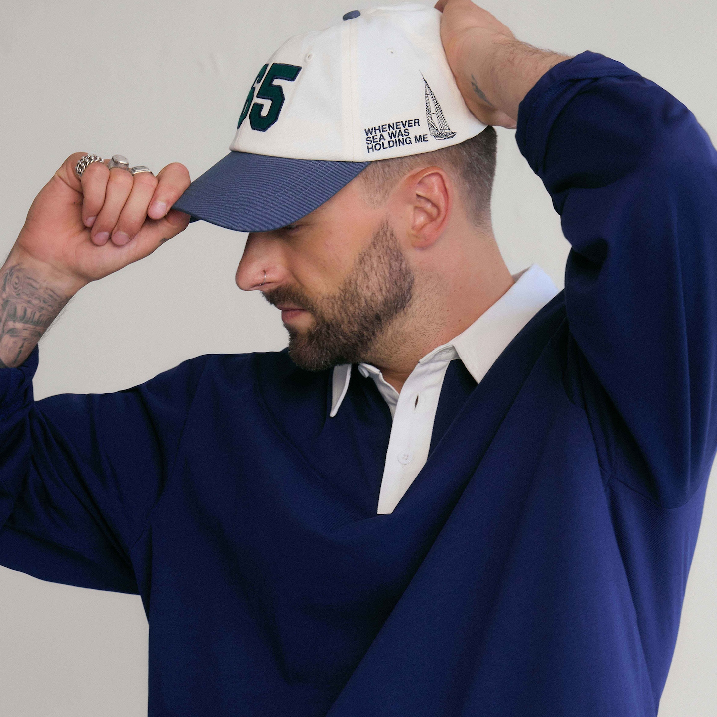 Model is wearing a navy long sleeve rugby polo top with a white and blue cap.