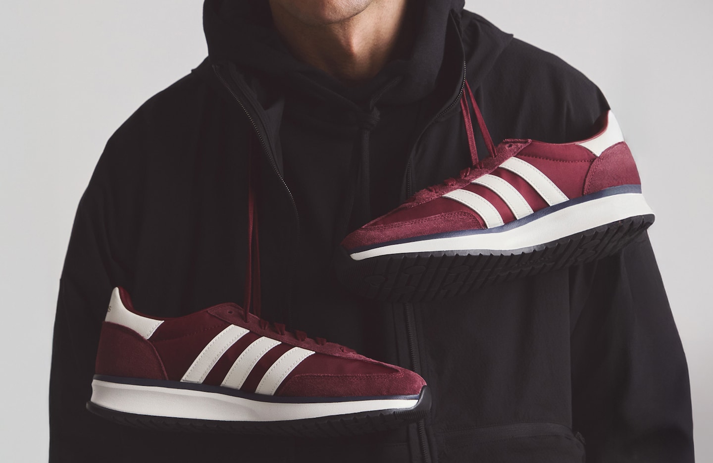 Model is wearing a black adidas Myshelter wind cheater jacket with a pair of dark red adidas Run 70s 2.0 trainers around his neck