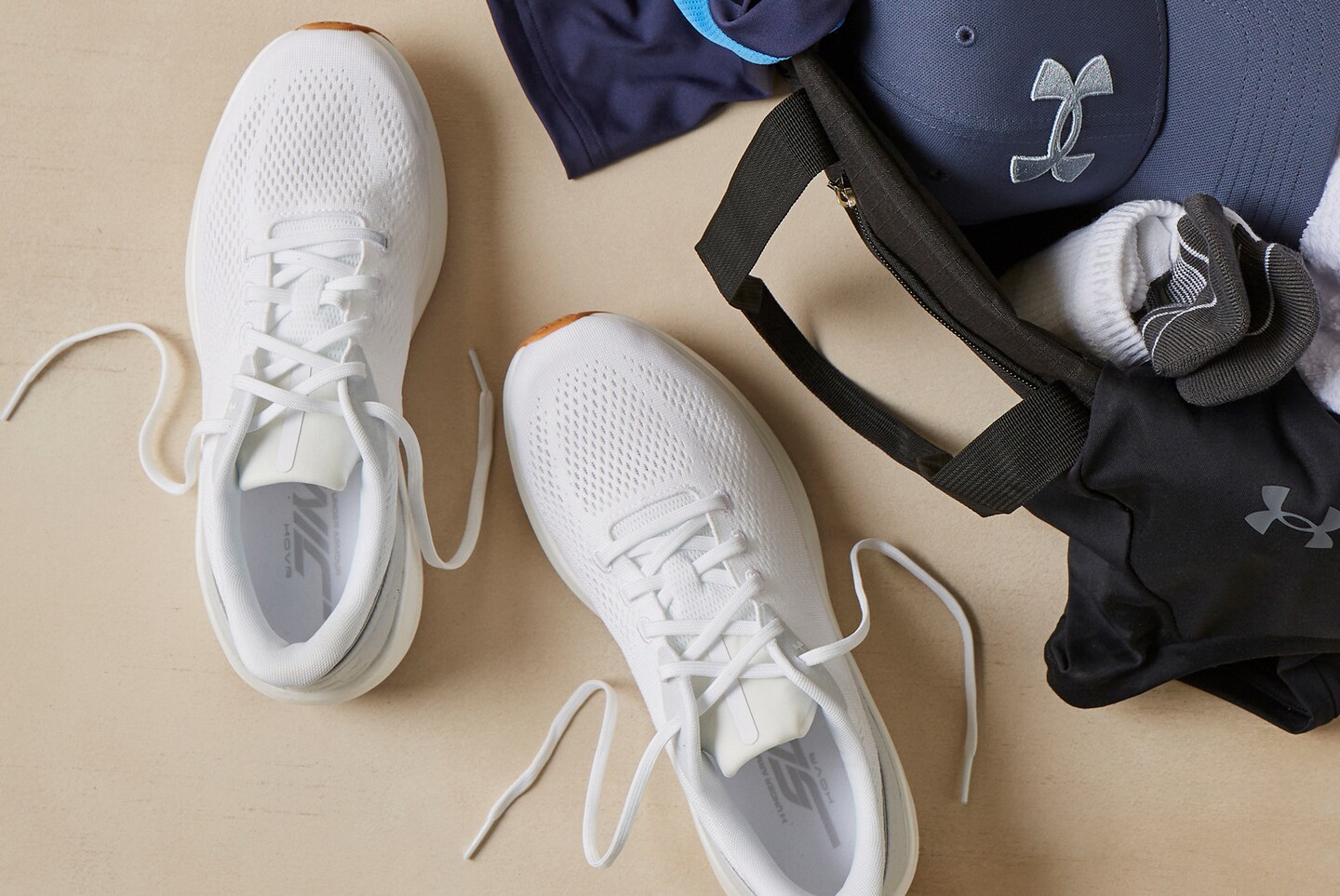 Pair of white trainers placed next to a black gym bag containing clothes and a grey cap with an Under Armour logo.