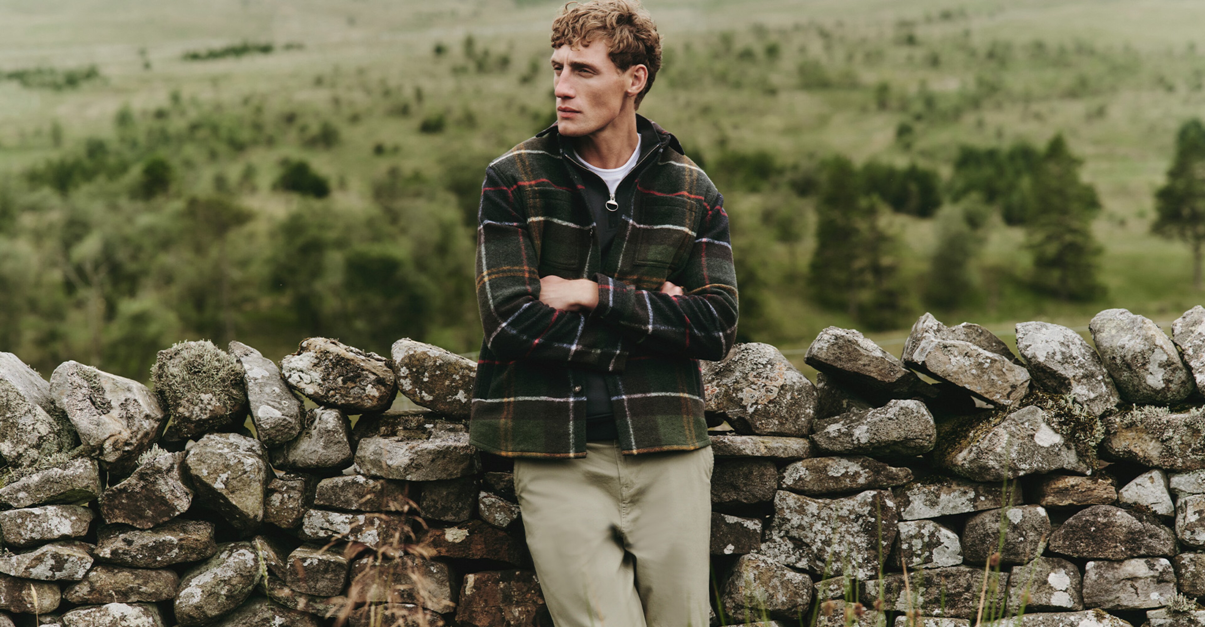 Model in the countryside, leaning against a brick wall wearing a chequered Barbour jacket