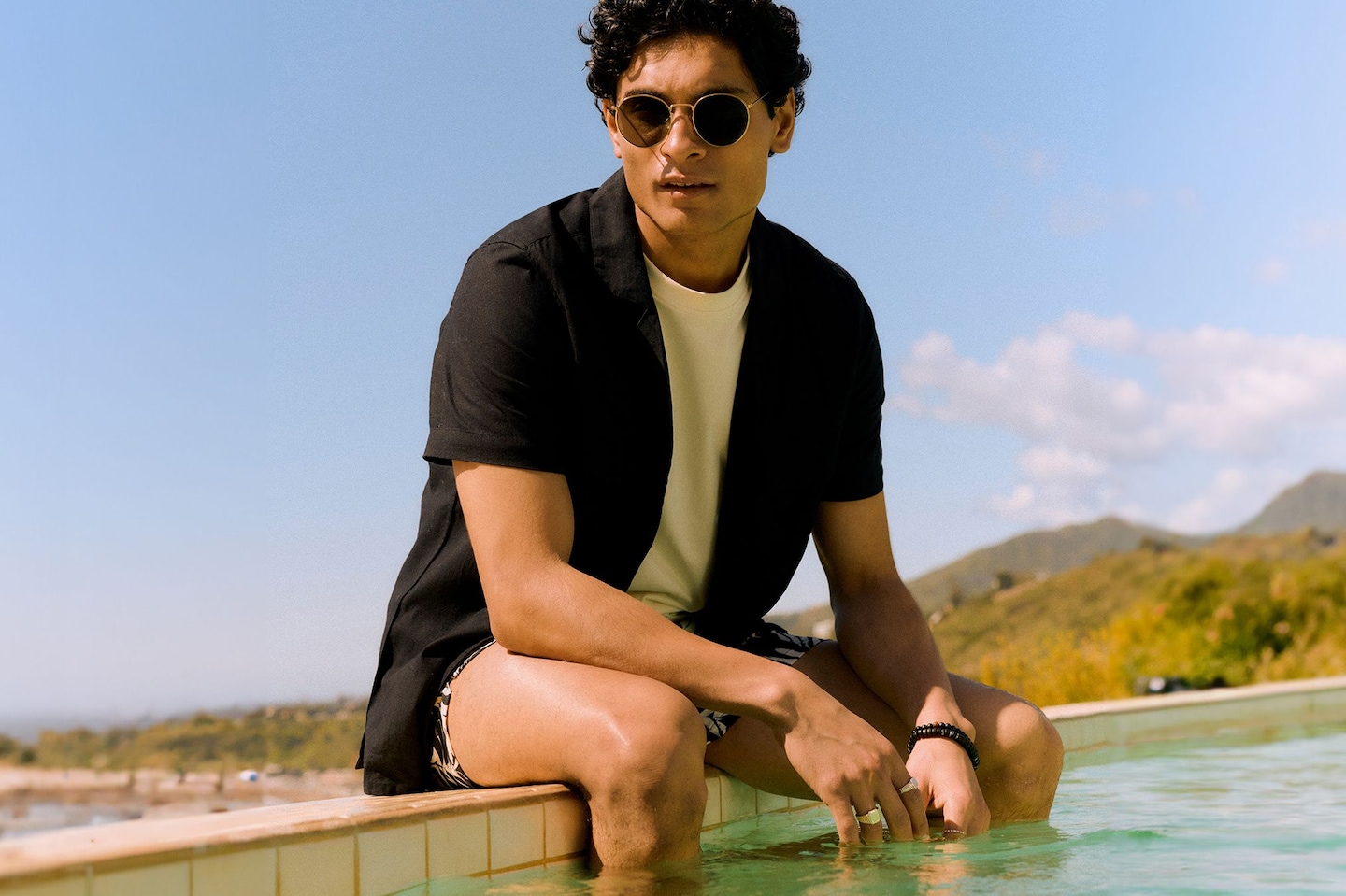 Model sat by the pool wearing black shirt, white t-shirt and swim shorts
