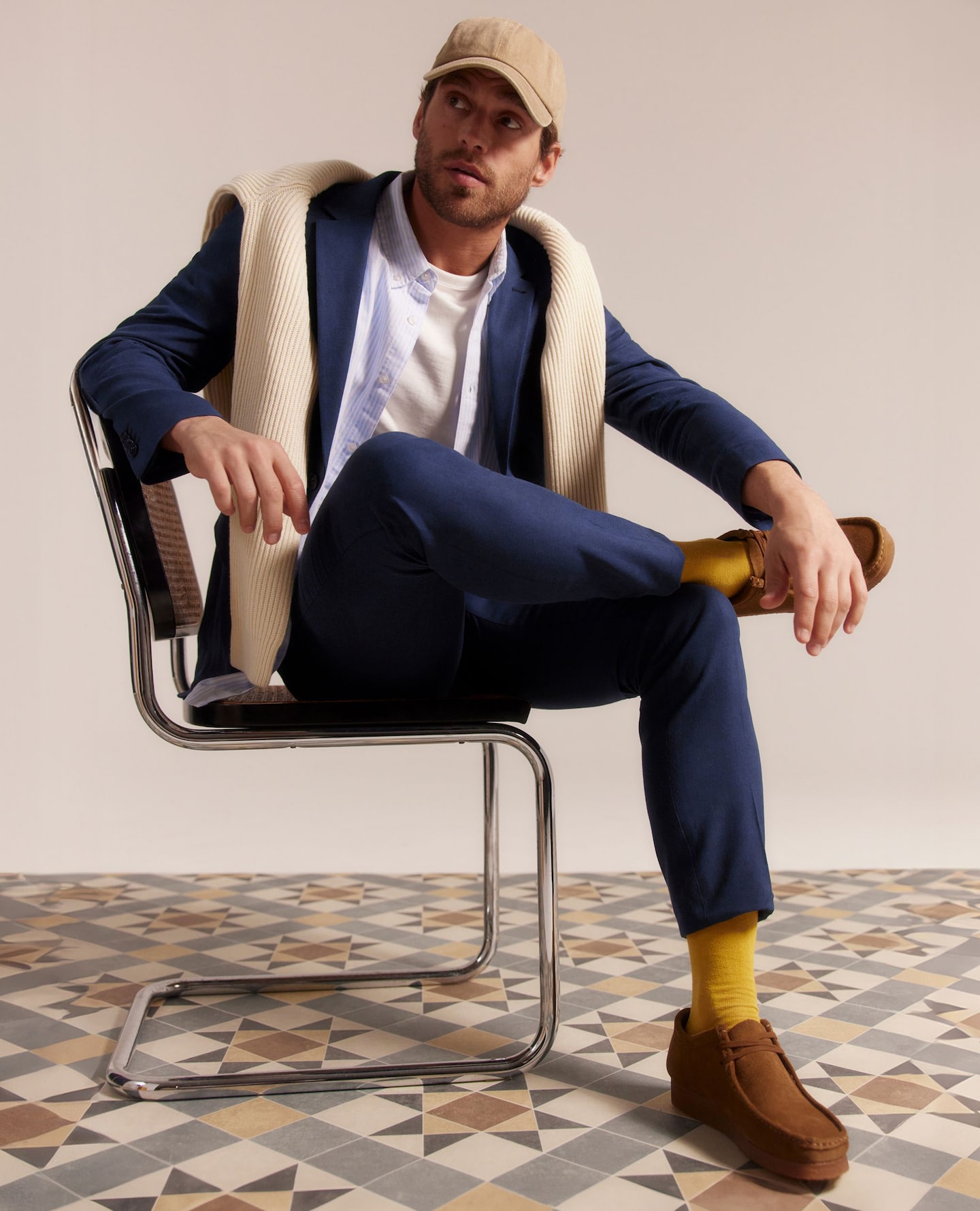 Model wears navy suit, blue shirt with white t-shirt with beige jumper and cap - also wears brown shoes.
