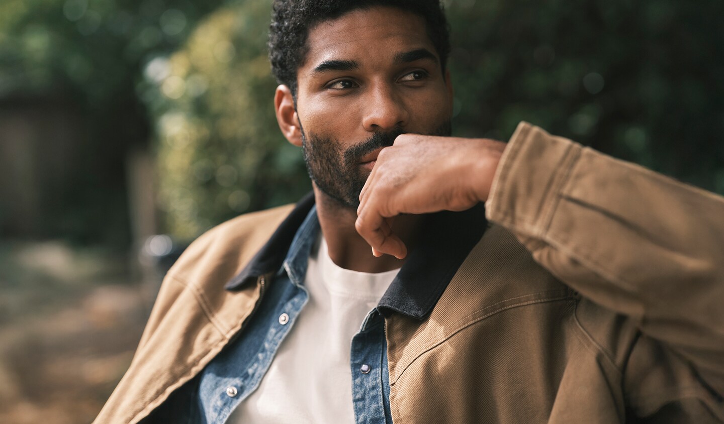 Model wears denim shirt and beige jacket, sat in a park setting