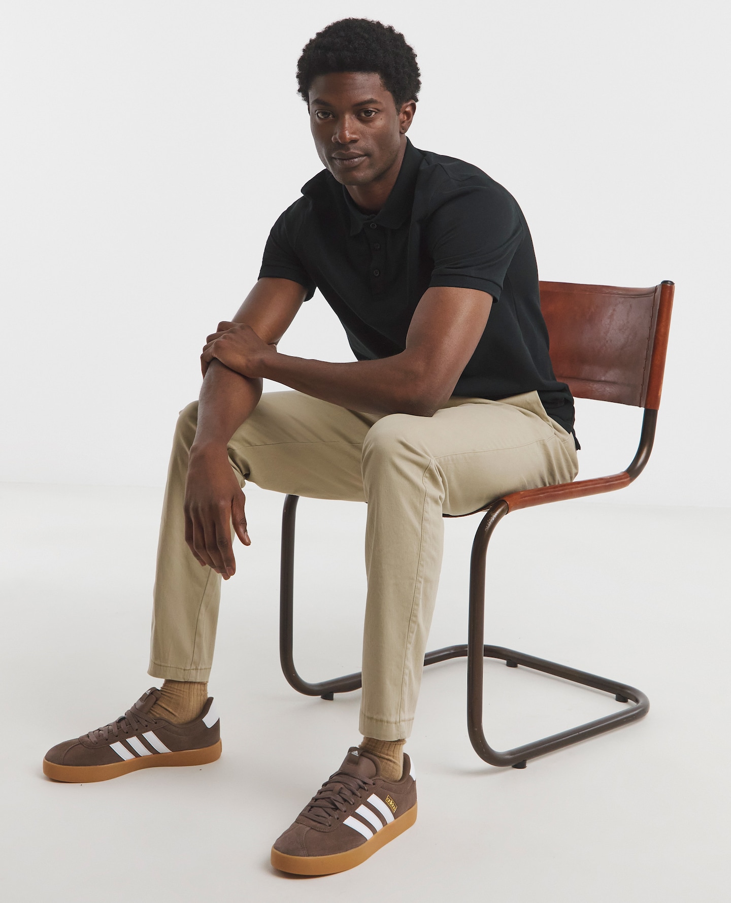 Model sat down wearing adidas trainer, cream chinos and a black polo