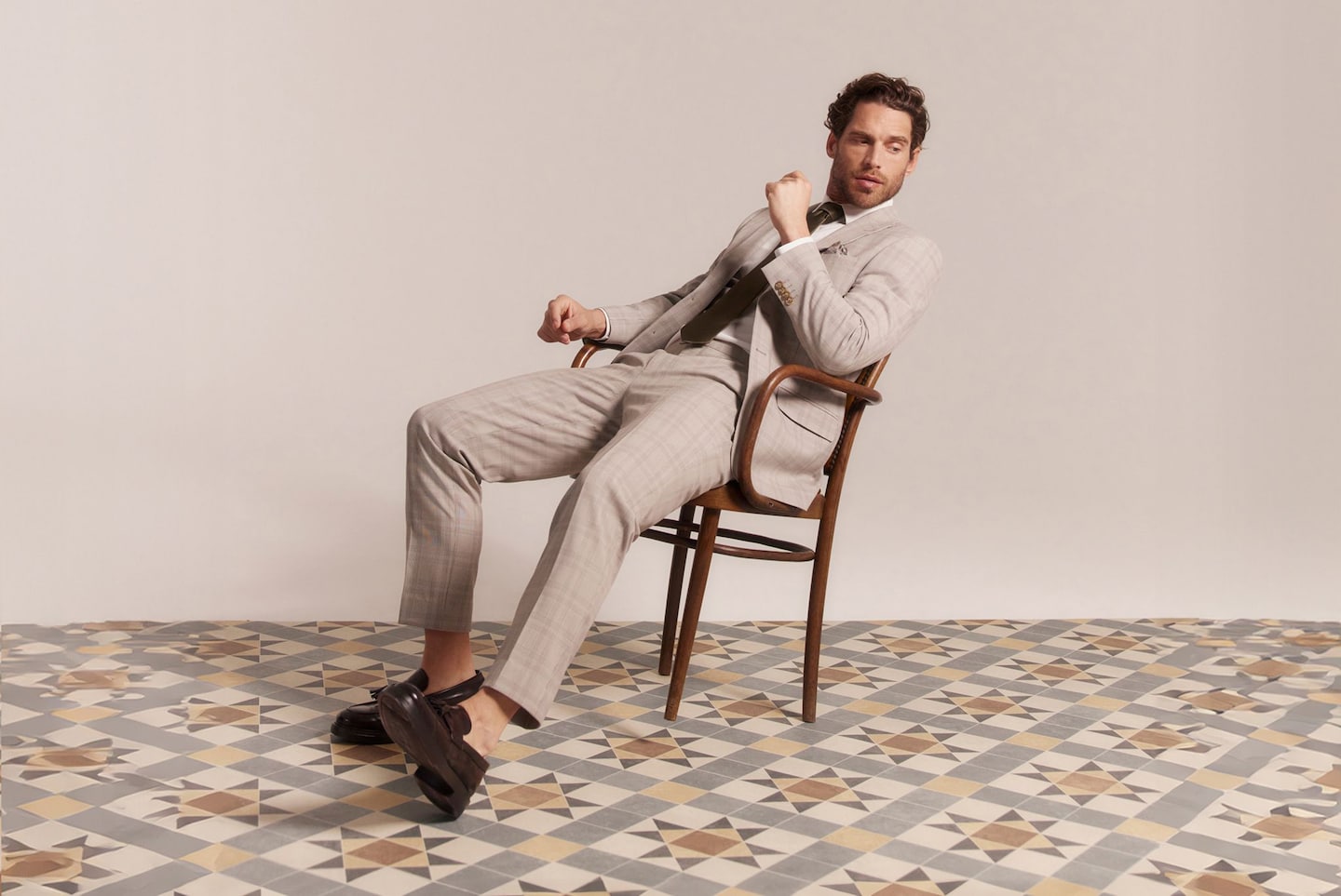 Model sat on chair wears smart beige suit, brown tie and brown smart shoes